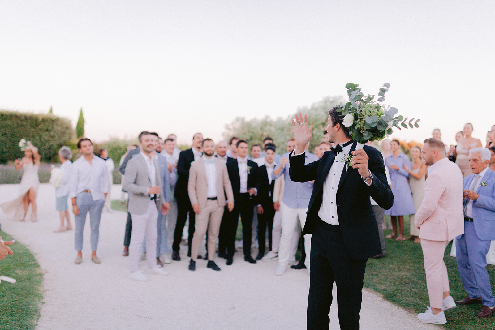 idee originale de lancer son bouquet, le marié tient dans la main sa boutonnière et s'apprête à la lancer derrière lui avec les invités hommes dans son dos